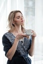 Beautiful young blonde woman in a blue robe by the window. Drinks coffee or tea from a white cup with a saucer. Morning Royalty Free Stock Photo