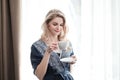 Beautiful young blonde woman in a blue robe by the window. Drinks coffee or tea from a white cup with a saucer. Morning Royalty Free Stock Photo