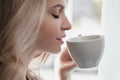 Beautiful young blonde woman in a blue robe by the window. Drinks coffee or tea from a white cup with a saucer. Morning Royalty Free Stock Photo