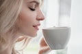 Beautiful young blonde woman in a blue robe by the window. Drinks coffee or tea from a white cup with a saucer. Morning Royalty Free Stock Photo