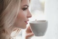 Beautiful young blonde woman in a blue robe by the window. Drinks coffee or tea from a white cup with a saucer. Morning Royalty Free Stock Photo
