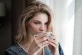 Beautiful young blonde woman in a blue robe by the window. Drinks coffee or tea from a white cup with a saucer. Morning Royalty Free Stock Photo