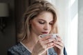 Beautiful young blonde woman in a blue robe by the window. Drinks coffee or tea from a white cup with a saucer. Morning Royalty Free Stock Photo