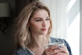 Beautiful young blonde woman in a blue robe by the window. Drinks coffee or tea from a white cup with a saucer. Morning Royalty Free Stock Photo