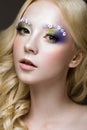 Beautiful young blonde woman with creative make-up color, curls and flowers on eyebrows. Beauty face. Art makeup. Royalty Free Stock Photo