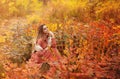 Beautiful young blonde woman in dress sitting with pensive look in tall grass in autumn forest