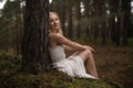 Beautiful young blonde woman sitting in forest nymph in white dress in evergreen wood Royalty Free Stock Photo