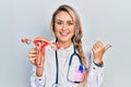 Beautiful young blonde woman holding anatomical model of female genital organ pointing thumb up to the side smiling happy with Royalty Free Stock Photo