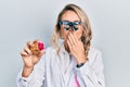 Beautiful young blonde woman holding geode stone wearing magnifier glasses covering mouth with hand, shocked and afraid for