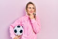 Beautiful young blonde woman holding soccer ball serious face thinking about question with hand on chin, thoughtful about Royalty Free Stock Photo