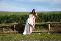 A beautiful young blonde woman with long hair in a wedding dress fastens her high heels while sitting on a wooden fence. In the Royalty Free Stock Photo