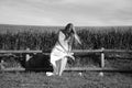 A beautiful young blonde woman with long hair in a wedding dress fastens her high heels while sitting on a wooden fence. In the Royalty Free Stock Photo