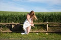 A beautiful young blonde woman with long hair in a wedding dress fastens her high heels while sitting on a wooden fence. In the Royalty Free Stock Photo