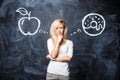 Beautiful young blonde woman makes a choice between an apple and a donut on the background of a chalk board. Healthy eating