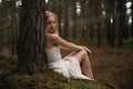 Beautiful young blonde woman sitting in forest nymph in white dress in evergreen wood Royalty Free Stock Photo