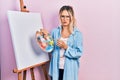 Beautiful young blonde woman standing drawing with palette by painter easel stand skeptic and nervous, frowning upset because of