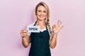 Beautiful young blonde woman wearing waitress apron holding open banner doing ok sign with fingers, smiling friendly gesturing Royalty Free Stock Photo