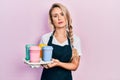 Beautiful young blonde woman wearing waitress apron holding take away cup of coffees relaxed with serious expression on face Royalty Free Stock Photo