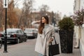 Beautiful young brown-haired woman in a sweater in a fashionable long light trench coat with a black leather bag posing on a