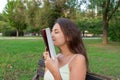 Beautiful young brownhair girl smells a fresh printed book sitting on the bench in the park