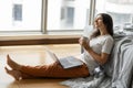 Beautiful young brunette girl working on a laptop and drinking coffee, sitting on the floor near the bed by the panoramic window. Royalty Free Stock Photo