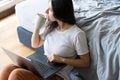 Beautiful young brunette girl working on a laptop and drinking coffee, sitting on the floor near the bed. Stylish modern interior Royalty Free Stock Photo