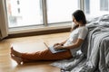 Beautiful young brunette girl working on a laptop, sitting on the floor near the bed by the panoramic window. Stylish modern Royalty Free Stock Photo