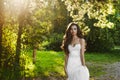 Beautiful and young brunette model girl, in white lacy dress, is posing in the park at the sunset