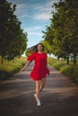 Beautiful young brunette in a red summer dress with flowing brown hair walks along a bicycle path and in an avenue of trees on a