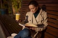 Beautiful young brunette sitting on wooden bench in park, reading book and drinking coffee. Serene confident woman in green Royalty Free Stock Photo