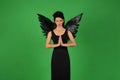 Beautiful young brunette woman in black angel costume with wings over green background in praying pose. Halloween party Royalty Free Stock Photo