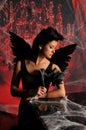 Beautiful young brunette woman in black angel costume with wings over spooky red background posing by piano with rose Royalty Free Stock Photo