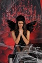 Beautiful young brunette woman in black angel costume with wings over spooky red background posing by piano with rose