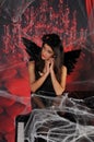 Beautiful young brunette woman in black angel costume with wings over spooky red background posing by piano with rose