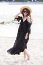 Beautiful young brunette woman in black dress, black sunglasses and straw hat walking on the sand on the beach and Royalty Free Stock Photo