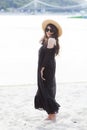 Beautiful young brunette woman in black dress, black sunglasses and straw hat walking on the sand on the beach and Royalty Free Stock Photo