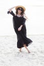Beautiful young brunette woman in black dress, black sunglasses and straw hat walking on the sand on the beach and Royalty Free Stock Photo