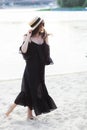 Beautiful young brunette woman in black dress, black sunglasses and straw hat walking on the sand on the beach and Royalty Free Stock Photo