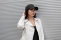 Beautiful young brunette woman in a fashionable white leather jacket in a T-shirt in a stylish black baseball cap posing Royalty Free Stock Photo