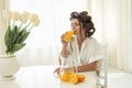 Beautiful young brunette woman in hair curlers enjoying orange fresh squeezed juice drinking it from glass sitting in Royalty Free Stock Photo