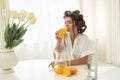 Beautiful young brunette woman in hair curlers enjoying orange fresh squeezed juice drinking it from glass sitting in bright Royalty Free Stock Photo