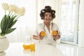 Beautiful young brunette woman in hair curlers holding oranges in both hands near her breasts in bright kitchen looking Royalty Free Stock Photo
