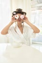 Beautiful young brunette woman in hair curlers holding two cupcakes in both hands near her eyes sitting with cup of coffee in Royalty Free Stock Photo