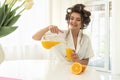 Beautiful young brunette woman in hair curlers pouring orange fresh squeezed juice from pitcher in glass sitting in Royalty Free Stock Photo