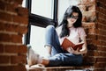 Beautiful young brunette woman reading book near window at home.