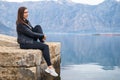 beautiful young brunette woman sits on stone pier and gazes thoughtfully out to sea, beautiful mountain range on Royalty Free Stock Photo