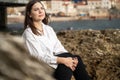 beautiful young brunette woman sits on stone pier and gazes thoughtfully out to sea, beautiful mountain range on Royalty Free Stock Photo