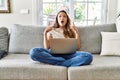Beautiful young brunette woman sitting on the sofa using computer laptop at home amazed and surprised looking up and pointing with Royalty Free Stock Photo