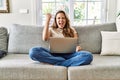 Beautiful young brunette woman sitting on the sofa using computer laptop at home angry and mad raising fist frustrated and furious Royalty Free Stock Photo