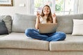 Beautiful young brunette woman sitting on the sofa using computer laptop at home gesturing finger crossed smiling with hope and